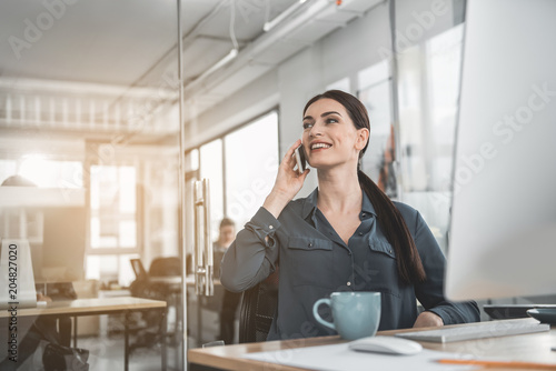 Portrait of beaming businesswoman talking by phone while working with computer at table