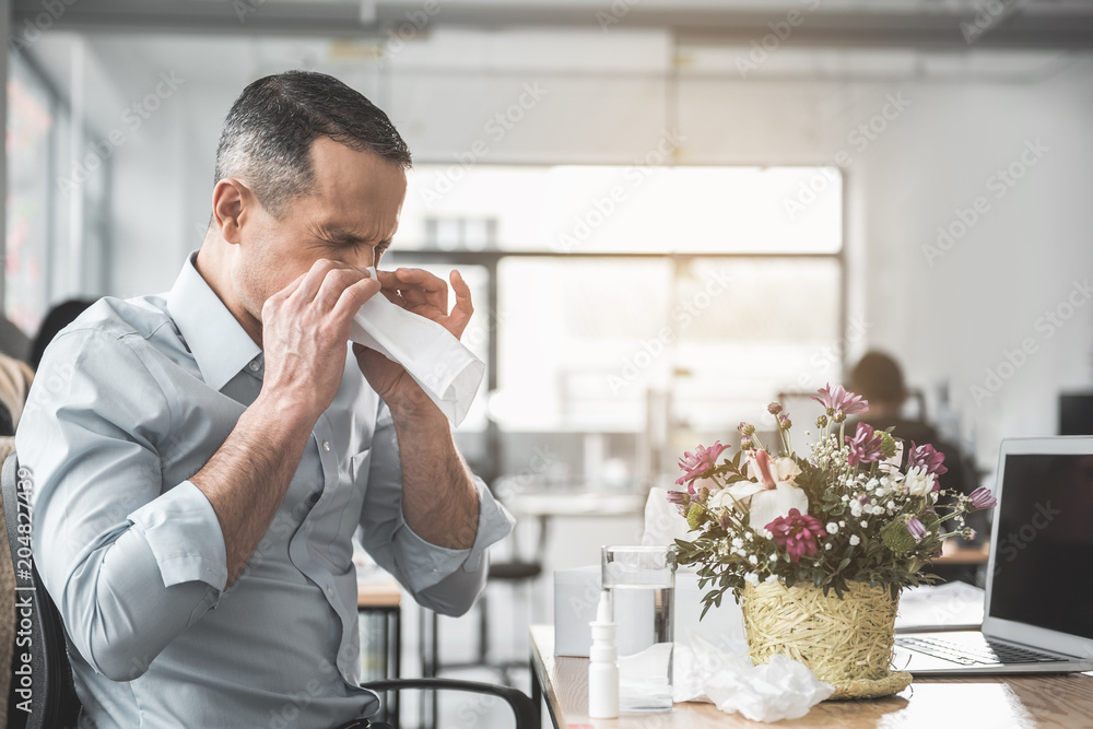 Side view unhappy ill businessman with running nose working with laptop in office. He sitting at table near pretty bouquet. Diseased employer at job concept Side view unhappy ill businessman with running nose working with laptop in office. He sitting at table near pretty bouquet. Diseased employer at job concept