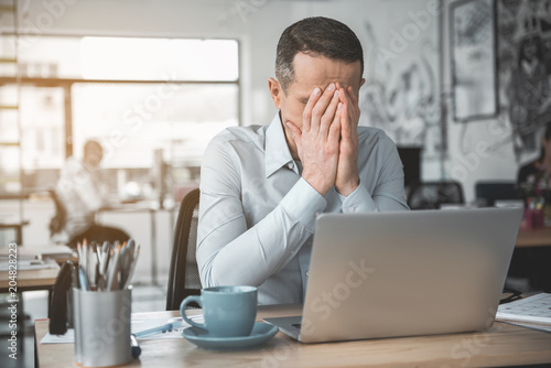 Depressed employer closing face while sitting at desk during job. Unhappy manager concept photo