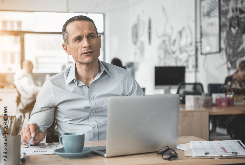 Pensive businessman reading document while typing in notebook computer. He locating at table. Wistful worker at job concept Pensive businessman reading document while typing in notebook computer. He locating at table. Wistful worker at job concept