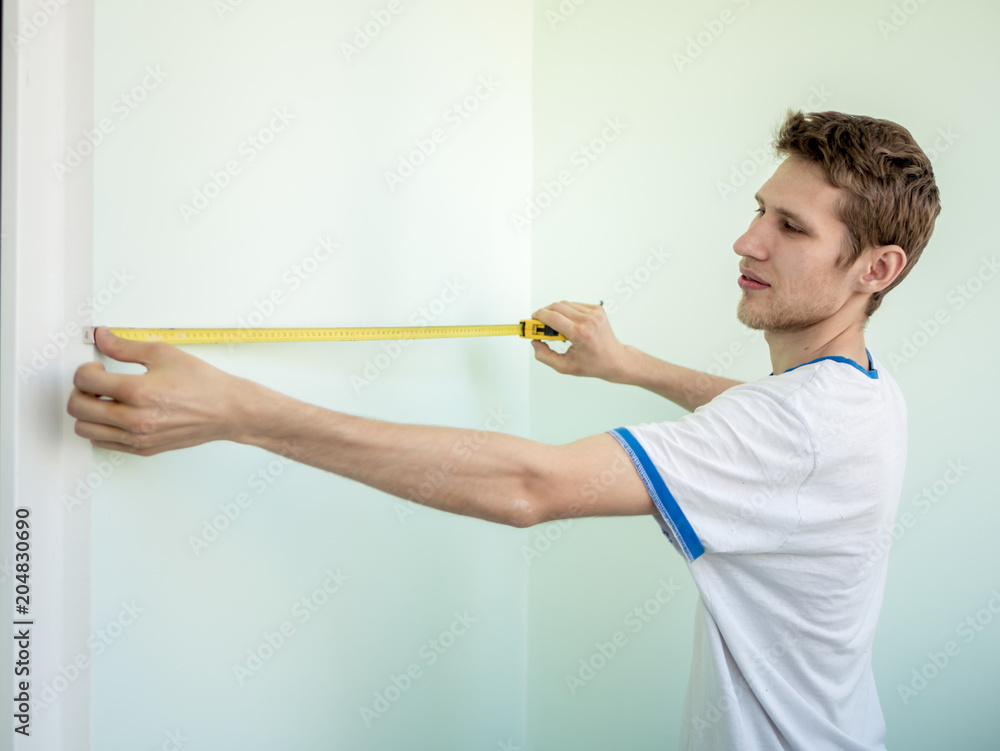 young man working at home measuring distance on the wall with roulette young man working at home measuring distance on the wall with roulette