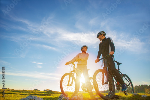 Cyclist couple with mountain bikes standing on the hill under the evening sky and enjoying bright sun at the sunset.