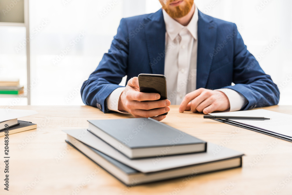 partial view of businessman in suit using smartphone at workplace with notebooks in office partial view of businessman in suit using smartphone at workplace with notebooks in office