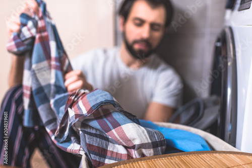 handsome loner taking clothes for washing near washing machine in bathroom with shirt on foreground