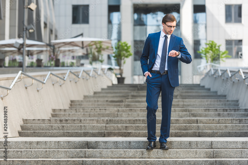 handsome young businessman in stylish suit standing on stairs near business building and looking at watch handsome young businessman in stylish suit standing on stairs near business building and looking at watch