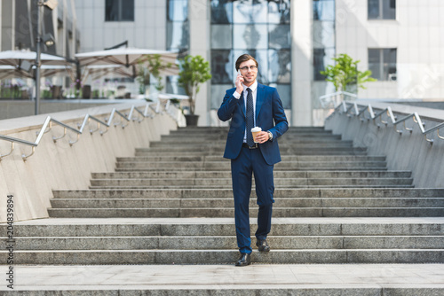 smiling young businessman in stylish suit with coffee to go talking by phone while walking by stairs near business building