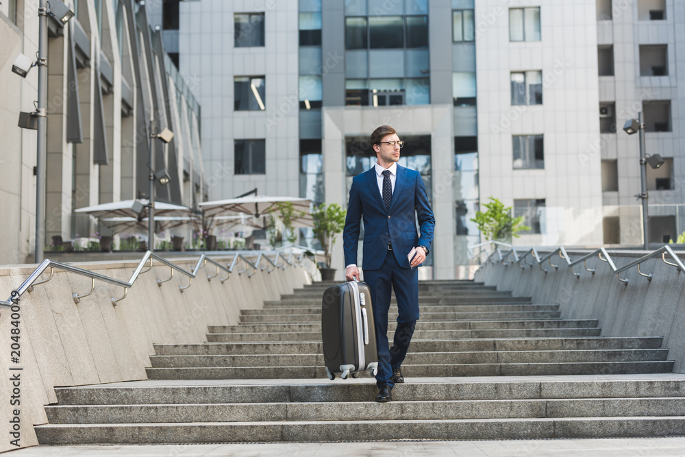 young businessman in stylish suit with luggage and flight tickets going down stairs in business district young businessman in stylish suit with luggage and flight tickets going down stairs in business district