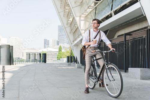 handsome young man with paper cup of coffee on vintage bicycle near sports stadium building