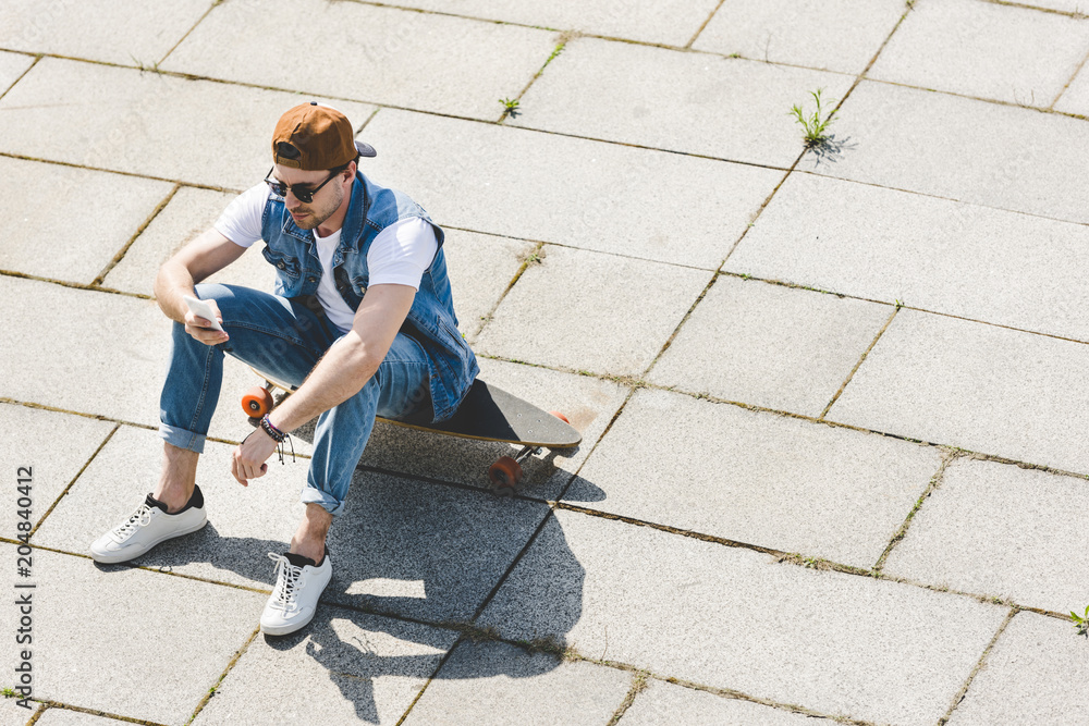 high angle view of stylish young man sitting on skateboard and using smartphone