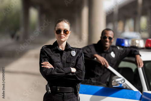Canvas Print young serious policewoman standing with crossed arms while her partner standing
