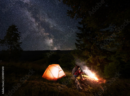 Young couple hikers sitting on a log, having a rest near burning campfire and illuminated orange tent under incredible beautiful starry sky with Milky way at night. Romantic night camping near forest