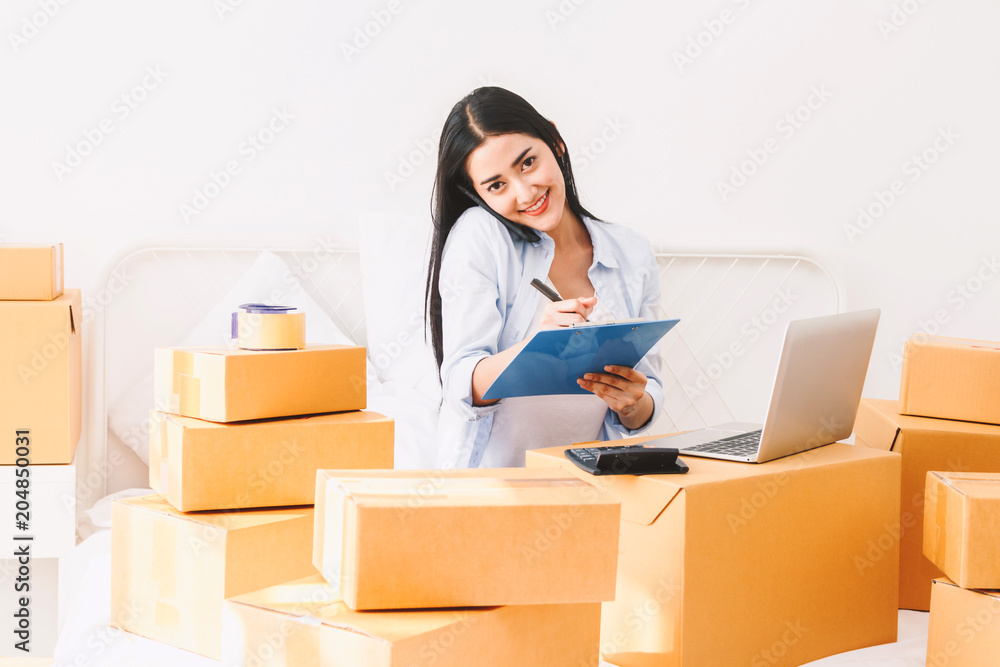 Young woman freelancer working and checklist and writing order with cardboard box on bed at home - SME business online and delivery concept Young woman freelancer working and checklist and writing order with cardboard box on bed at home - SME business online and delivery concept