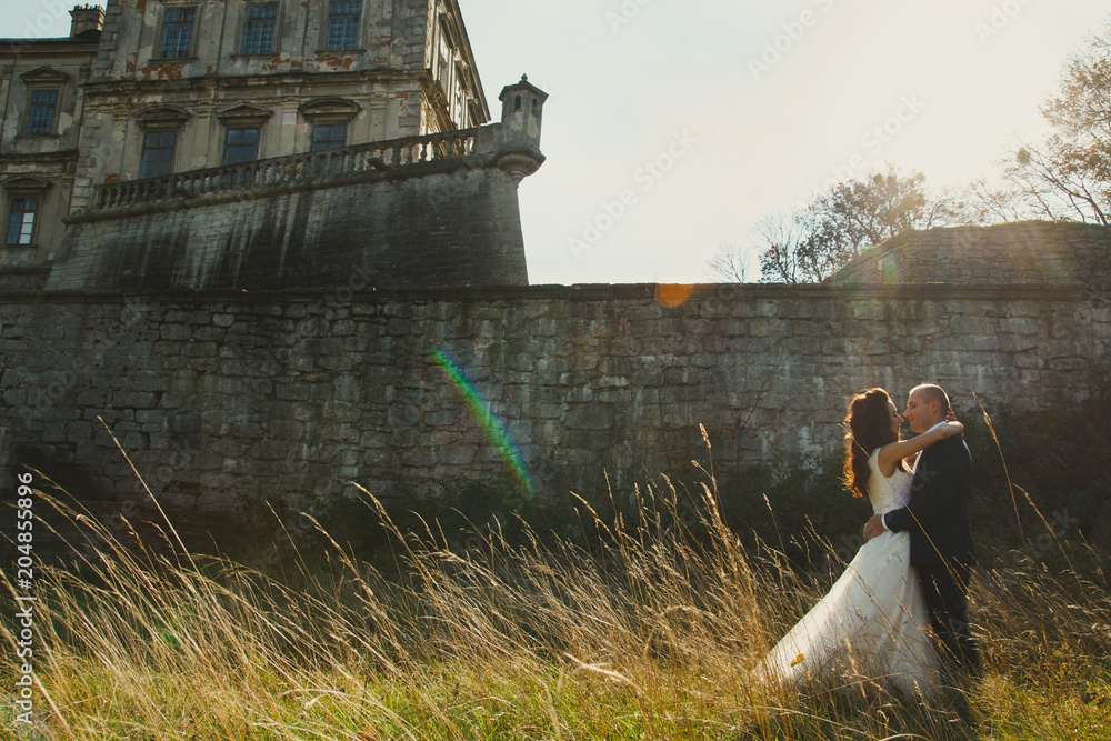 Obraz premium Wedding couple is walking outdoors in sunny day. Medieval stone walls with old renaissance palace on background. Bride is satin lace dress is embracing groom. Old architecture and dry high grass.