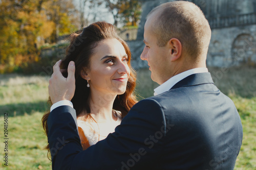 Wedding couple is walking outdoors in sunny day. Medieval stone walls with old renaissance palace on background. Groom touching bride's face in satin lace dress. Old architecture and green grass.