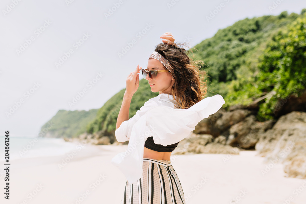 Back view of young pretty lovely woman with wavy dark hair dressed white shirt and striped pants turns around at camera on background of green rocks on the white beach near the ocean in sunlight