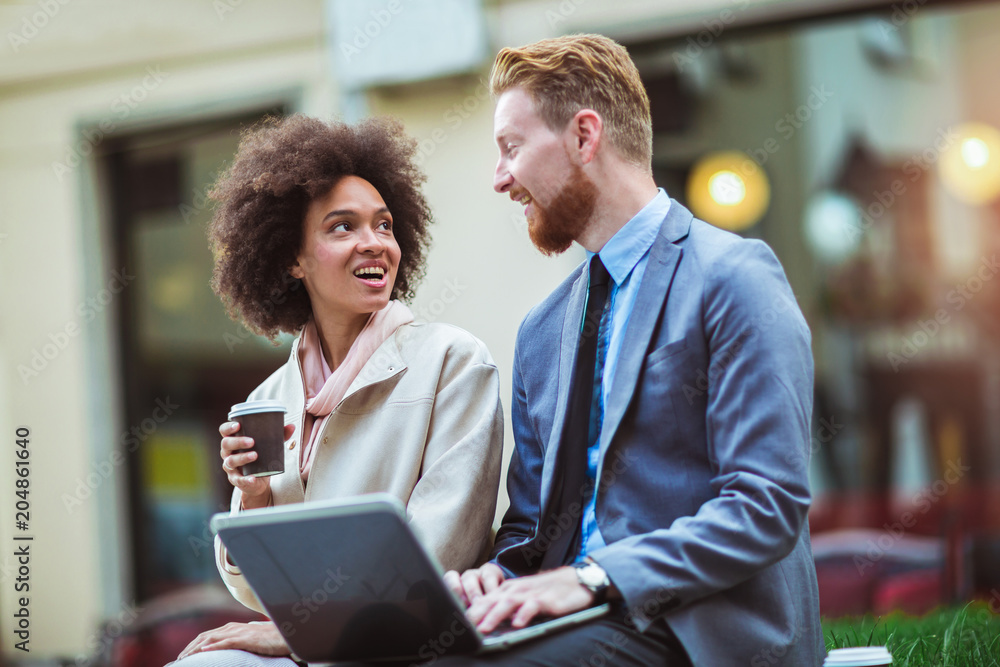 Two business people in an informal conversation in front of a business building using laptop and drink coffee Two business people in an informal conversation in front of a business building using laptop and drink coffee