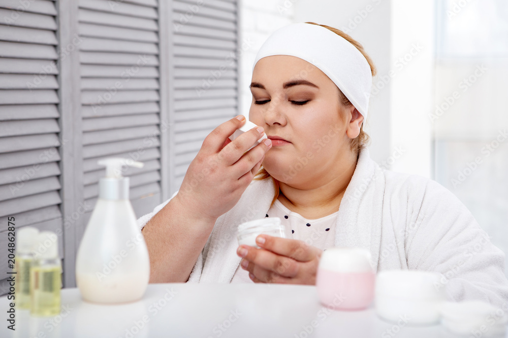 My skin care. Serious overweight woman wearing a bathrobe and smelling her skin cream My skin care. Serious overweight woman wearing a bathrobe and smelling her skin cream