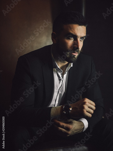 A handsome man with a beard and in a classic suit sits in a leather chair and looks at the camera. 