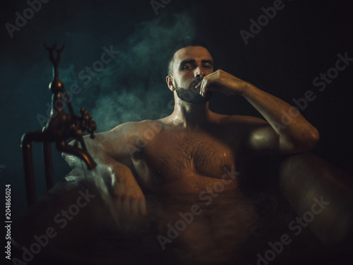 A handsome, muscular man with a beard lies in a bronze bathroom. Steam coming from the hot water. He looks at the camera, drops of water running down the body