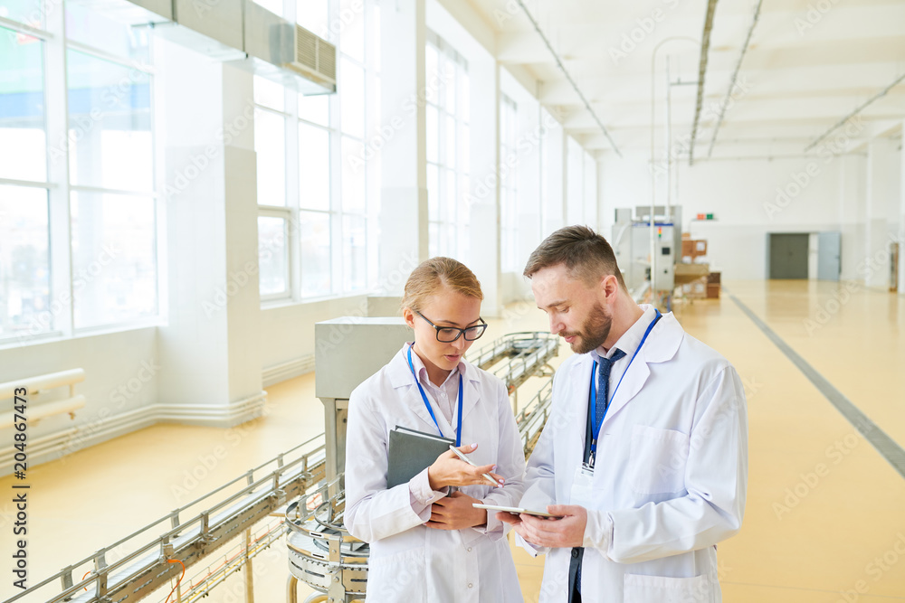 Fototapeta premium Confident inspectors wearing white coats using digital tablet while standing at spacious packaging department of milk factory