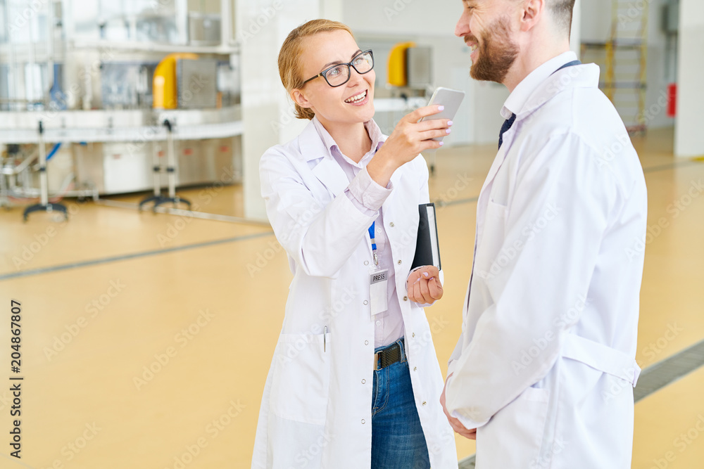 Smiling journalist wearing white coat using dictaphone on smartphone while talking to cheerful bearded owner of soy milk factory, interior of spacious production department on background Smiling journalist wearing white coat using dictaphone on smartphone while talking to cheerful bearded owner of soy milk factory, interior of spacious production department on background