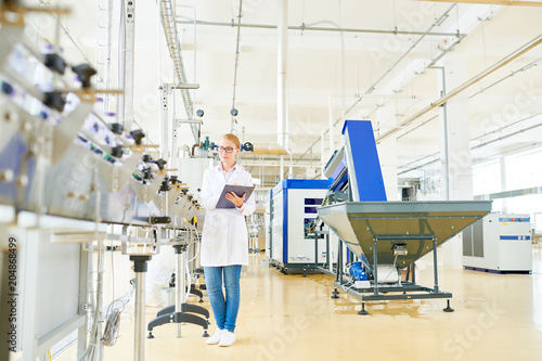 Concentrated fair-haired worker wearing white coat standing at conveyor belt and controlling production process at spacious dairy factory