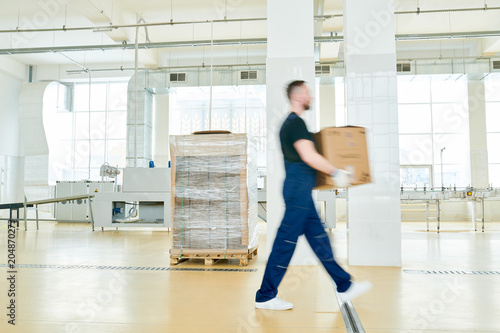 Motion shot of unrecognizable factory worker wearing overall carrying cardboard box, interior of packaging department on background