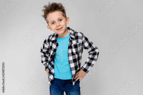 A handsome boy in a plaid shirt, blue shirt and jeans stands on a gray background. The boy put his hands on his hips