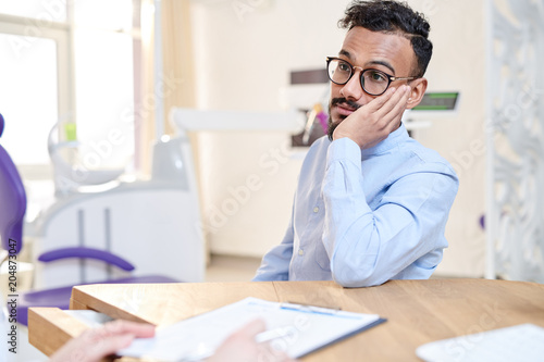 Portrait of worried  Middle-Eastern man suffering from toothache talking to doctor sitting at desk in modern dental clinic and describing symptoms