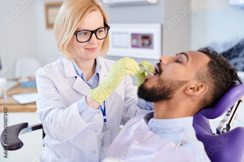 Portrait of smiling female dentist examining teeth of young patient sitting in dental chair