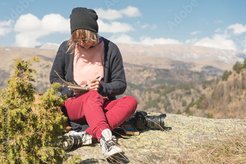 Portrait of a hipster girl wearing sunglasses and a hat sitting on a rock outdoor in the mountains against a blue sky. Freelancer the designer in travel draws a pencil in a notebook