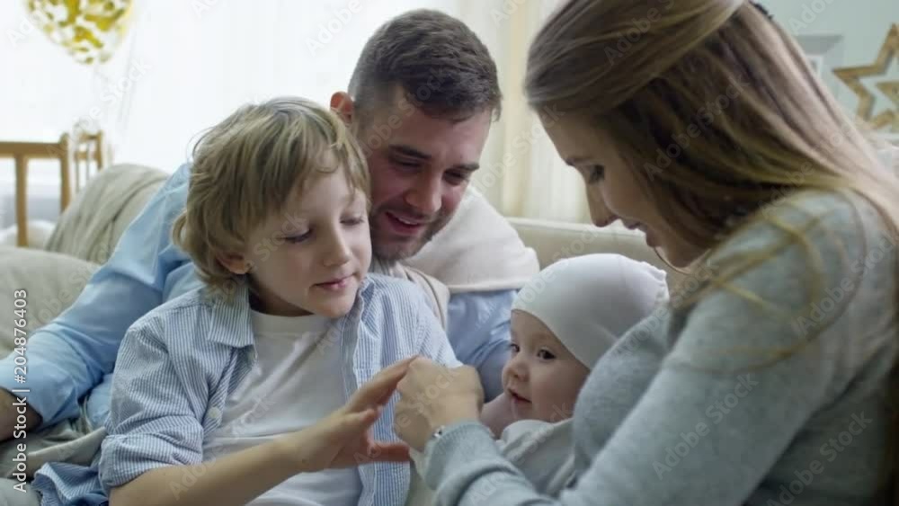 Tilt up of happy couple with elementary age boy sitting together on sofa and playing with baby boy Tilt up of happy couple with elementary age boy sitting together on sofa and playing with baby boy