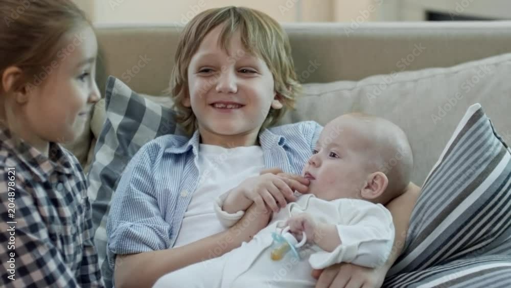 Medium shot of cute elementary age girl and boy sitting on sofa and playing with baby Medium shot of cute elementary age girl and boy sitting on sofa and playing with baby