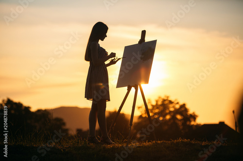 Silhouette of a blonde girl paints a painting on the canvas with the help of paints. A wooden easel keeps the picture. Summer is a sunny day, sunset