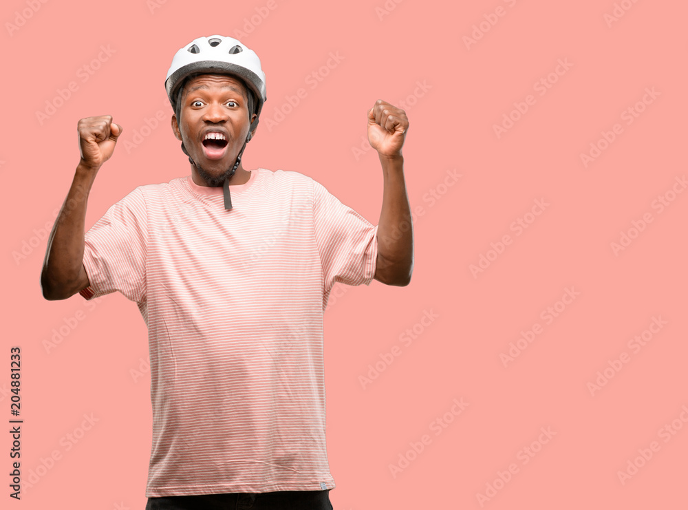 Black man wearing bike helmet happy and excited celebrating victory expressing big success, power, energy and positive emotions. Celebrates new job joyful