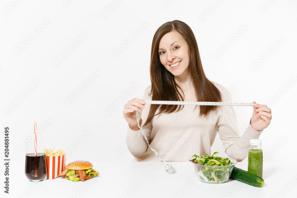 Woman with measure tape, green detox smoothies, salad in glass bowl, cucumber, burger, cola in bottle isolated on white background. Proper nutrition, healthy lifestyle, fast food, dieting concept.