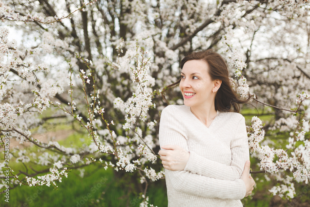 Young smiling happy charming woman in light casual clothes holding hands folded standing in city garden or park on blooming tree background. Spring nature, flowers. Lifestyle, leisure concept. Young smiling happy charming woman in light casual clothes holding hands folded standing in city garden or park on blooming tree background. Spring nature, flowers. Lifestyle, leisure concept.