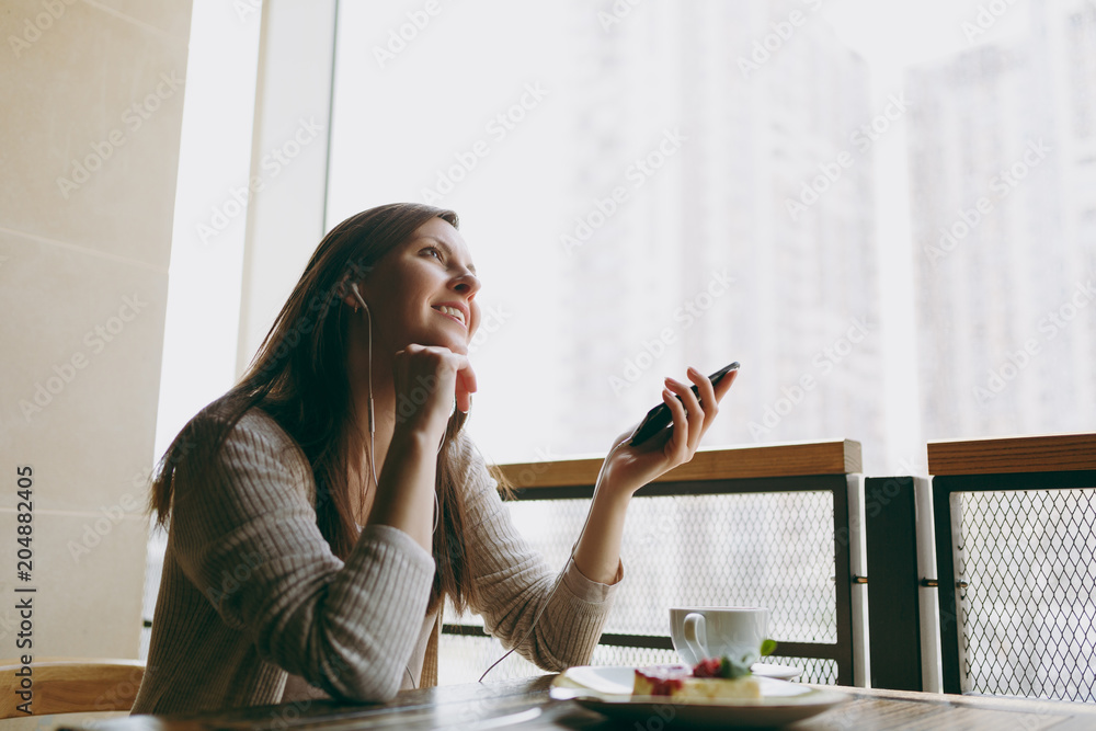 Young woman sitting in coffee shop at table with cup of cappuccino, cake, relax in restaurant during free time. Young female listen music in earphones on mobile phone, rest in cafe. Lifestyle concept. Young woman sitting in coffee shop at table with cup of cappuccino, cake, relax in restaurant during free time. Young female listen music in earphones on mobile phone, rest in cafe. Lifestyle concept.