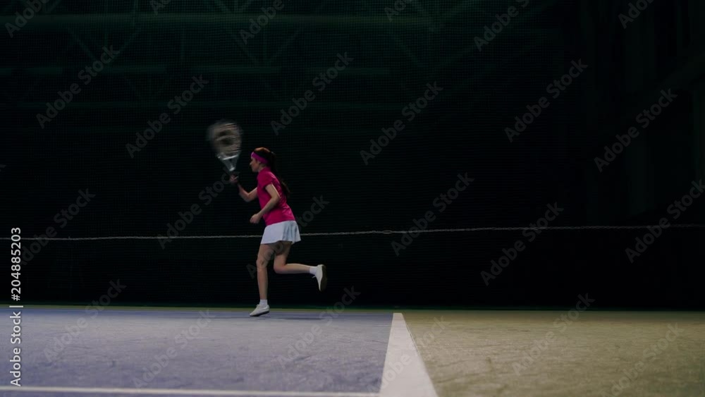 A woman in a pink t-shirt and a white skirt plays off the balls during a tennis match. The tennis player learns to play. Training tennis competition on a closed court in slow motion.