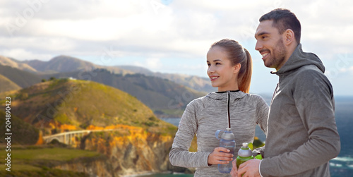 fitness, sport and people concept - smiling couple with bottles of water over bixby creek bridge on big sur coast of california background