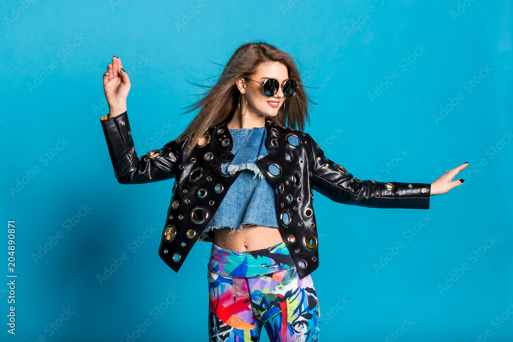 emotional stylish girl model in studio on a blue background, wearing bright leggings leather jacket sunglasses, dynamic pose, smile