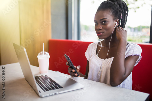Cheerful female black model resting in coffee shop listening favorite music in headphones and chatting in networks. Beautiful dark skinned freelancer working on laptop photo