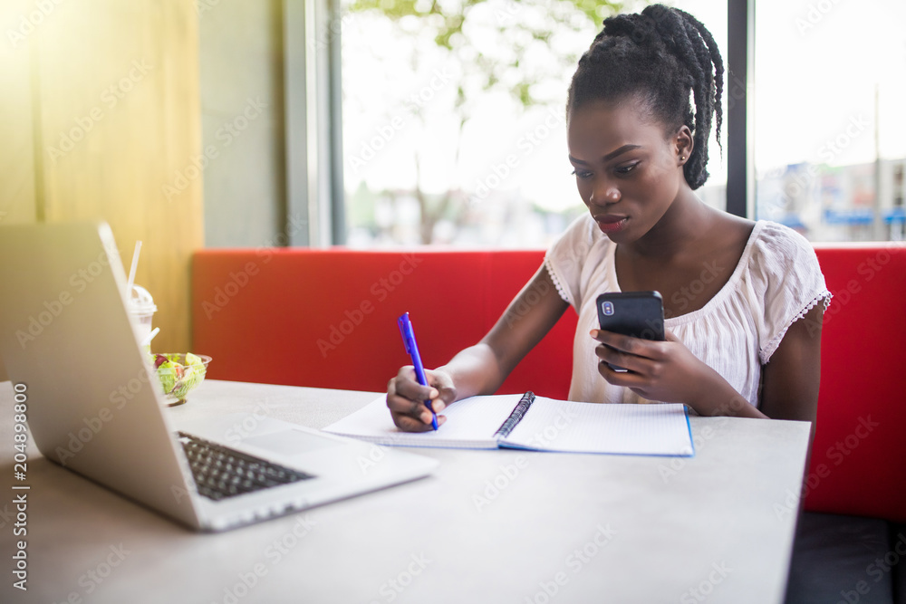 Fototapeta premium Young beautiful Afro-American woman using laptop and writing in notebook while studying in cafe