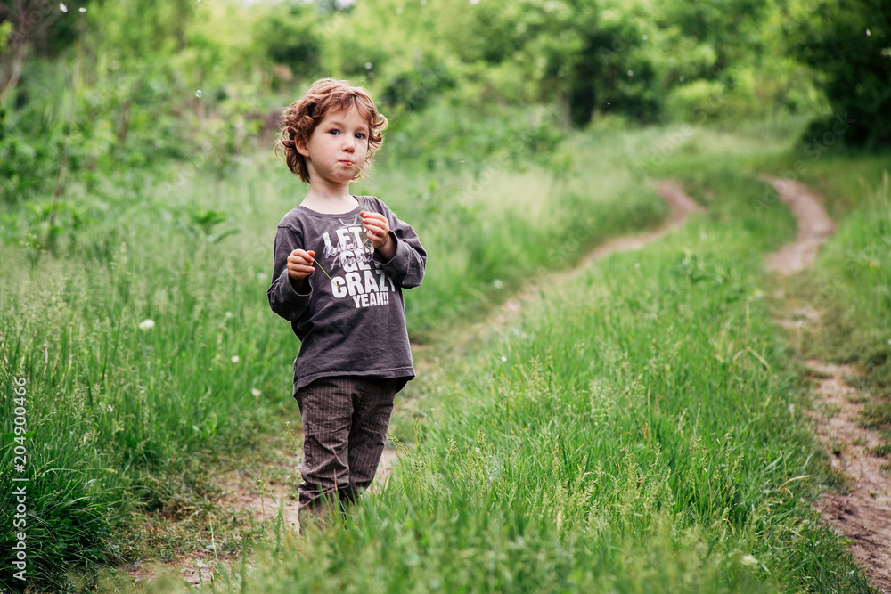 Adorable little toddler boy staying near the tree. Summer green grass, country road Adorable little toddler boy staying near the tree. Summer green grass, country road