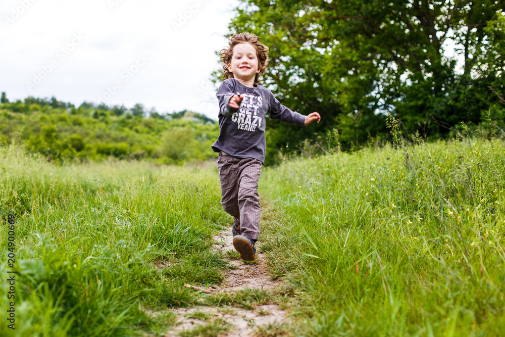 Little boy running down a meadow in a beautiful landscape in summer, very light and happy scene. Curly toddler Little boy running down a meadow in a beautiful landscape in summer, very light and happy scene. Curly toddler