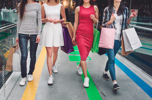 Cut view of girls walking together in a big store. They wear different clothes and have different colorful bags in their hands. Young women are on shopping.