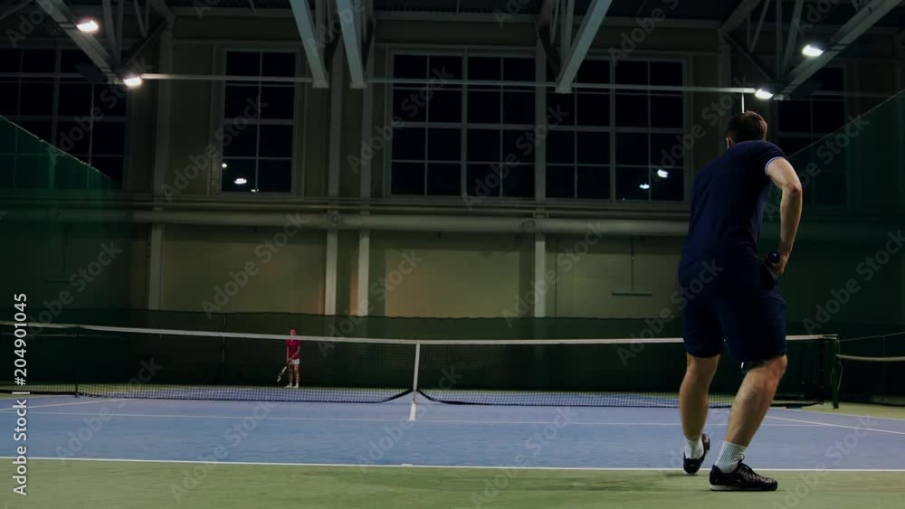 Serve by professional tennis player. Tennis serve indoor of tenis hall. Man on serve with tenis racket and dressed in black t-shirt, black shorts and black shoes. Serve by professional tennis player. Tennis serve indoor of tenis hall. Man on serve with tenis racket and dressed in black t-shirt, black shorts and black shoes.