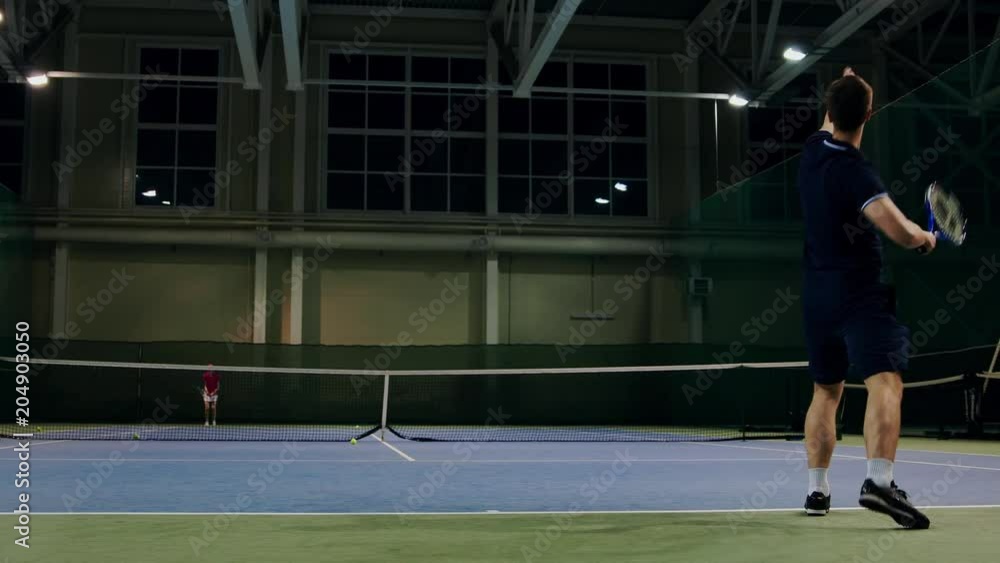 Serve by professional tennis player. Tennis serve indoor of tenis hall. Man on serve with tenis racket and dressed in black t-shirt, black shorts and black shoes. Serve by professional tennis player. Tennis serve indoor of tenis hall. Man on serve with tenis racket and dressed in black t-shirt, black shorts and black shoes.