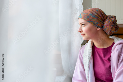 Young adult female cancer patient wearing headscarf and bathrobe sitting in the kitchen looking out window.