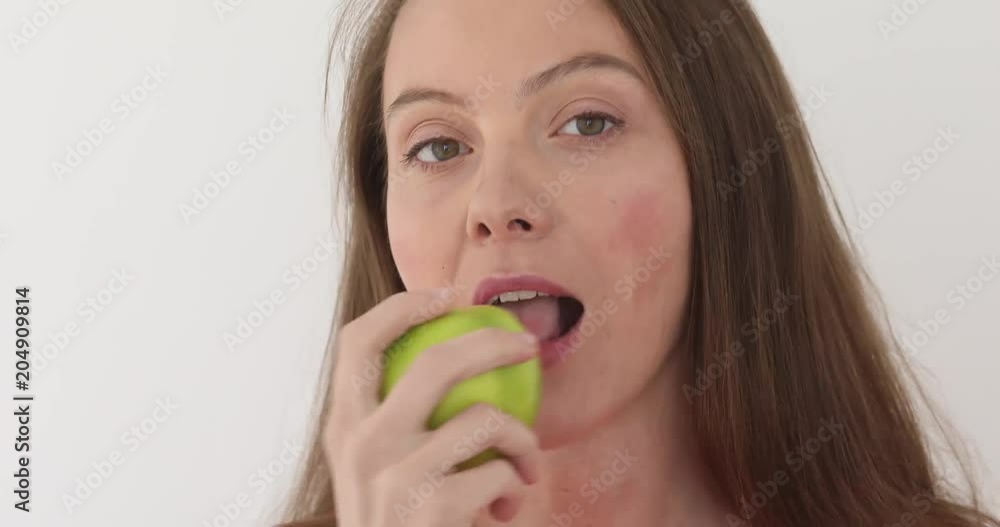 Young woman is eating a big green apple at white background. Healthy nutrition model eating fruit. Girl takes first bite and then offer bite to viewer and saying Wanna bite Young woman is eating a big green apple at white background. Healthy nutrition model eating fruit. Girl takes first bite and then offer bite to viewer and saying Wanna bite